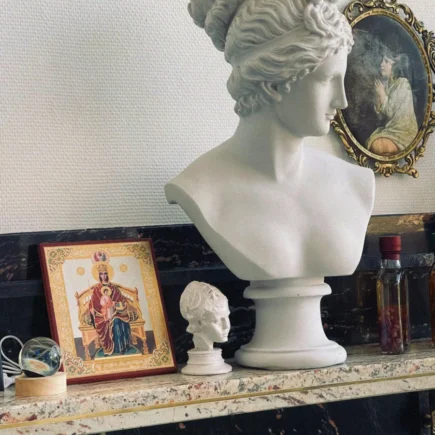 Marble bust of Venus alongside religious icon and antique mirror on ornate shelf.